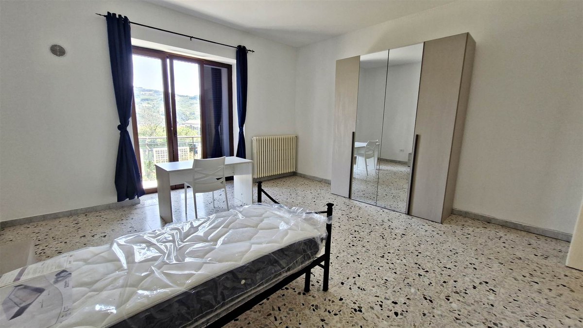 Camera con letto singolo e materasso imballato, scrivania bianca, armadio a specchi, tende blu e pavimento in graniglia